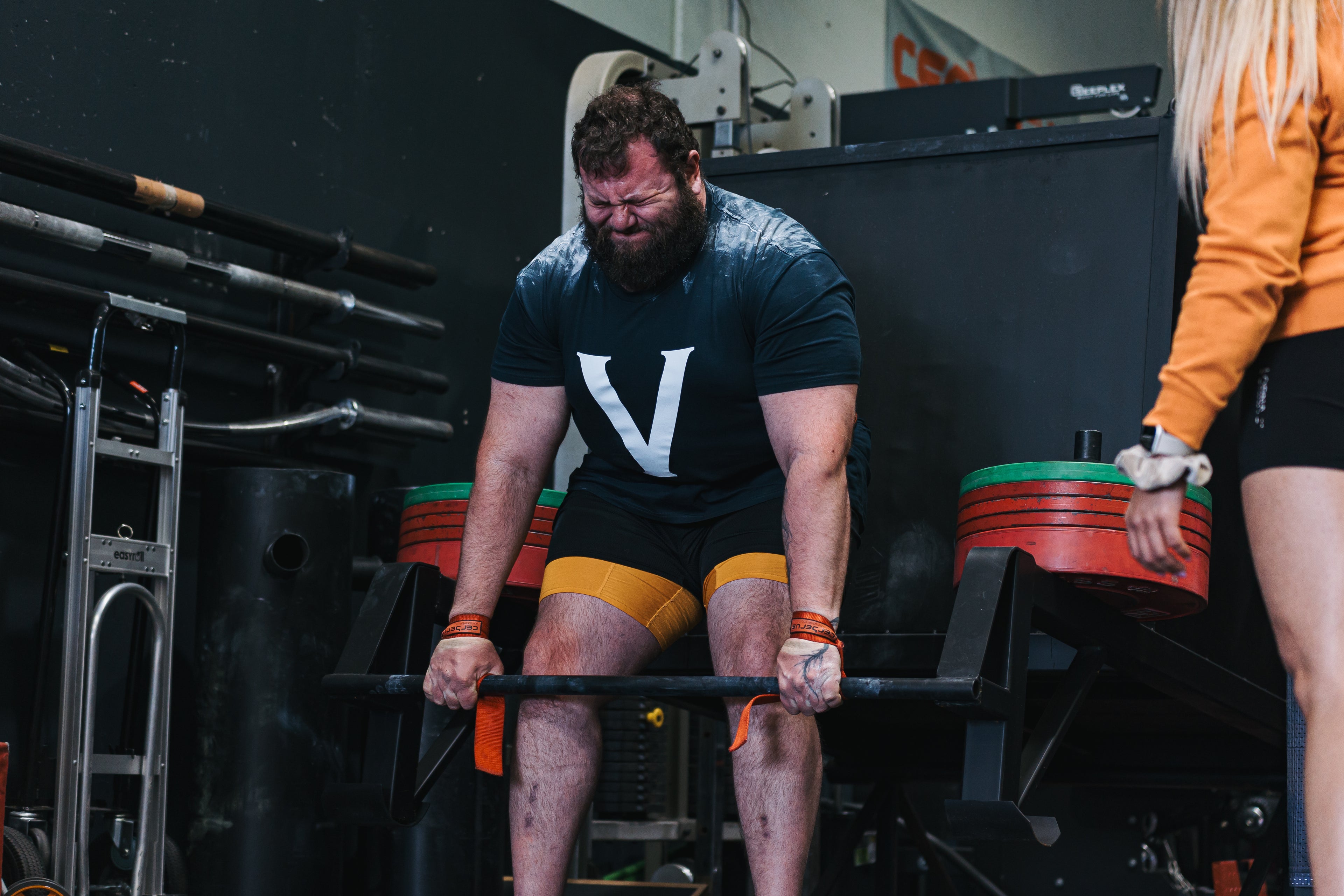 A bearded man in a gym wearing a black t-shirt with a large white letter 'V' and yellow compression sleeves, lifting a barbell on a rack. Another person in an orange sweatshirt is partially visible on the right.
