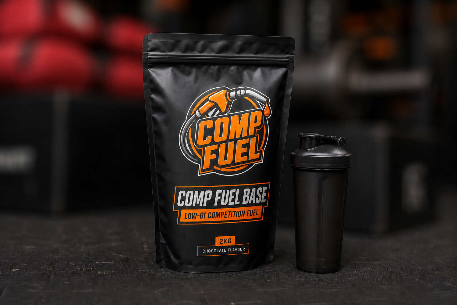 A black stand-up pouch of Comp Fuel Base chocolate flavor powder stands next to a matching black shaker bottle on a dark surface, with a blurred gym background.