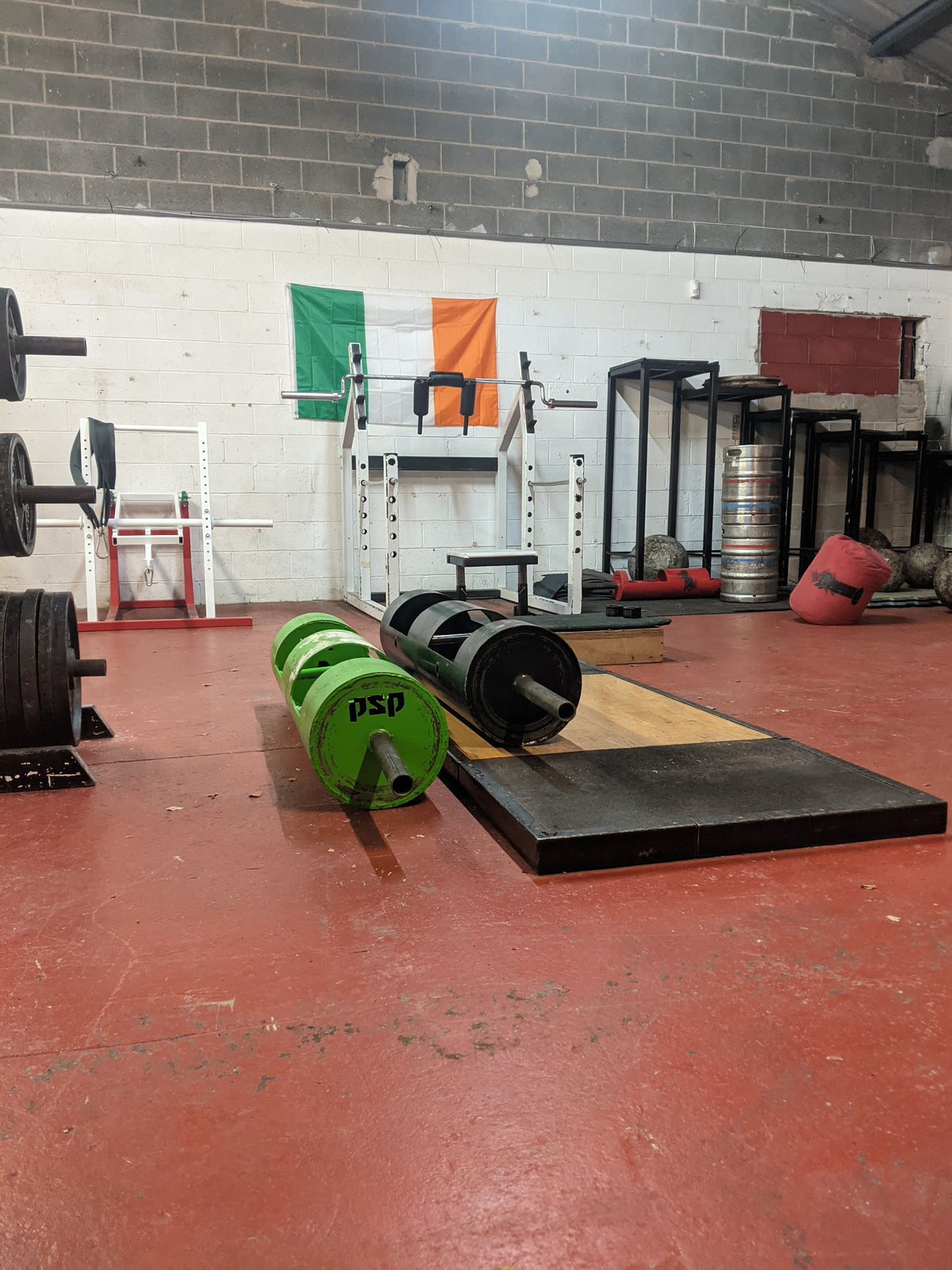 A barbell with bright green and black weight plates resting on a wooden weightlifting platform inside a gym with a red floor and brick walls.