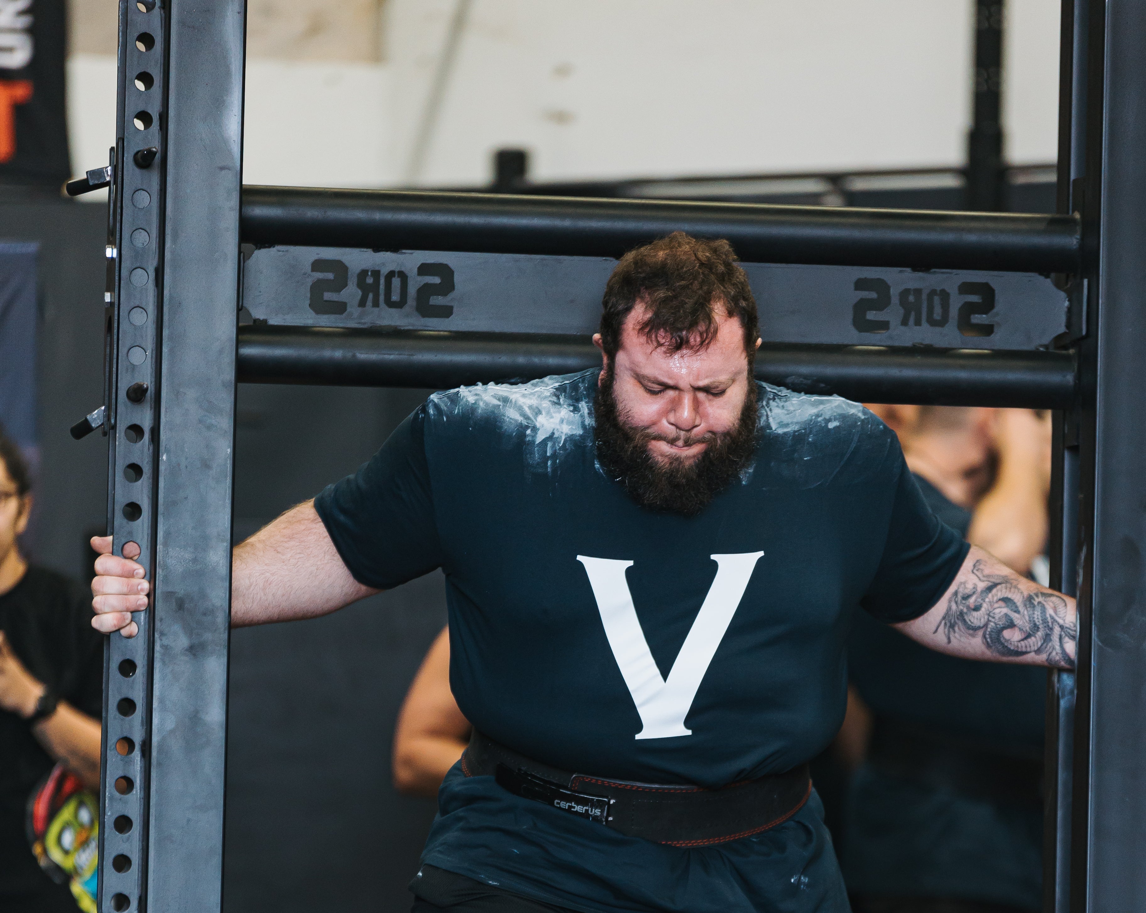 A bearded man in a gym wearing a dark navy t-shirt with a large white 'V' logo, black shorts, and bright orange knee sleeves, standing in front of a weight rack.