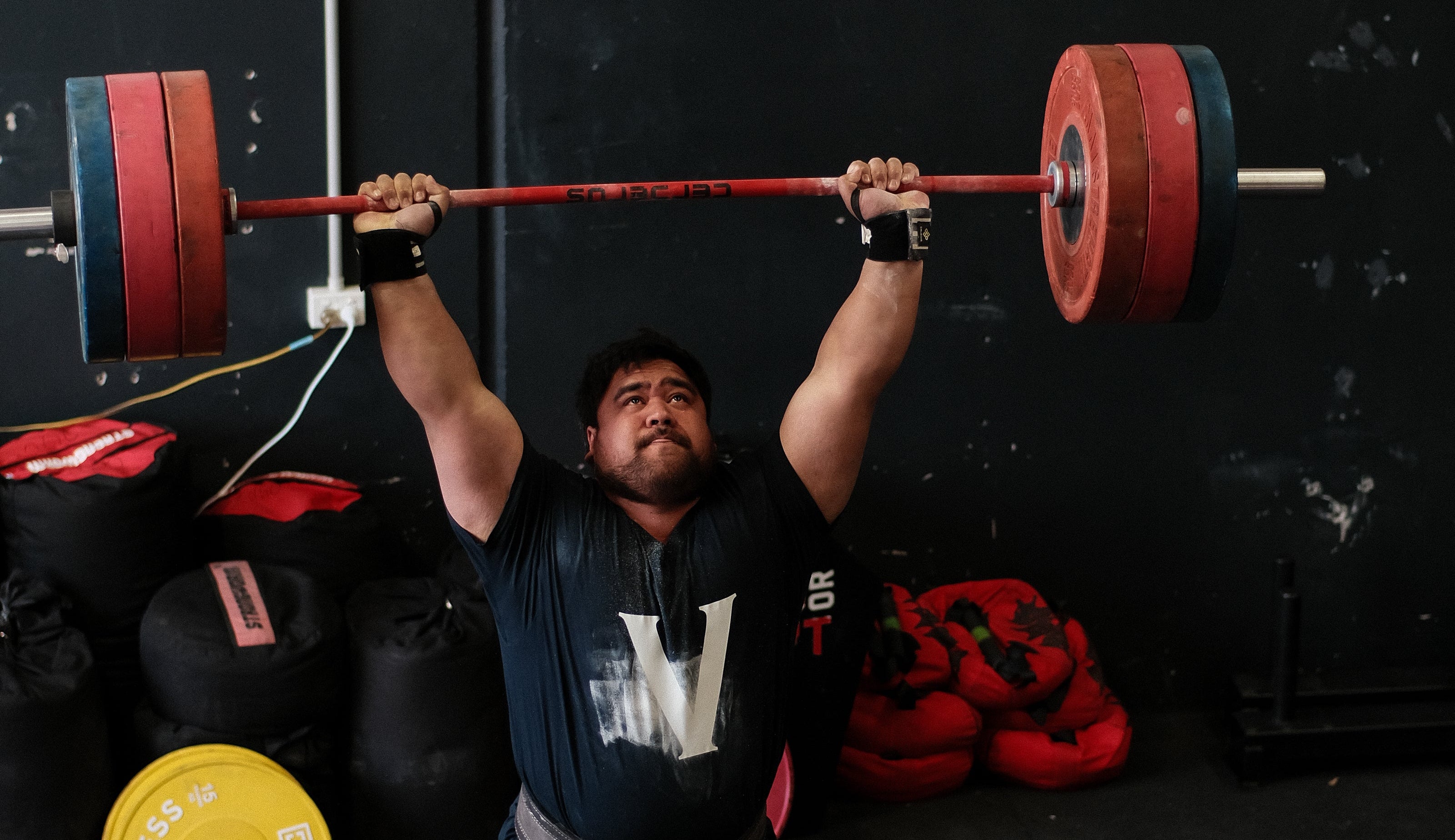 A man in a gym lifts a red barbell with red and blue weight plates overhead, wearing a black t-shirt, dark shorts, and weightlifting shoes. Sandbags and a yellow weight plate are visible in the background against a dark wall.